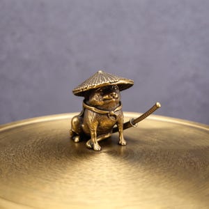 Peut inclure: Figurine de chien en bronze portant un chapeau de paille japonais traditionnel et une épée. Le chien est assis sur une surface circulaire dorée. La figurine présente des détails complexes et une esthétique vintage.