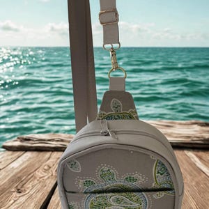 May include: A beige sling backpack featuring a repeating green and blue sea turtle pattern with paisley details. The bag has a single adjustable strap with gold-toned metal hardware, a top zipper closure, and a front pocket with a zipper.