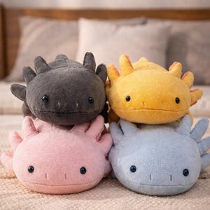 May include: Four plush axolotl toys in shades of grey, yellow, pink, and blue. Each has a rounded body, small black eyes, and a frilly head crest. They are stacked on a textured, light-coloured surface.
