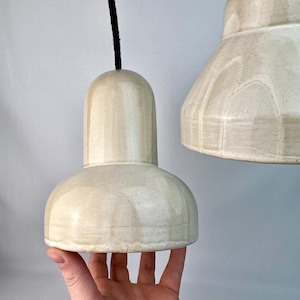 May include: Cream ceramic pendant lights with a speckled finish. The lights have a bell-shaped design, suspended by black cords. One light is held by a hand, showing the size and shape. The background is plain white.