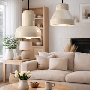 Puede incluir: Dos lámparas colgantes color crema cuelgan sobre un salón. Las luces tienen forma cilíndrica y cónica. La habitación cuenta con un sofá beige, una mesa de centro de madera y un jarrón con flores blancas, creando un ambiente cálido.