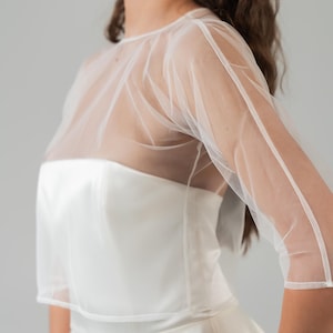 Op de afbeelding: Witte bruidsjurk met een doorschijnende, korte mouwen overlay. De jurk heeft een getailleerd lijfje en een zwierige rok. De overlay voegt een delicate touch toe aan het klassieke ontwerp, perfect voor een bruiloft of speciale gelegenheid.