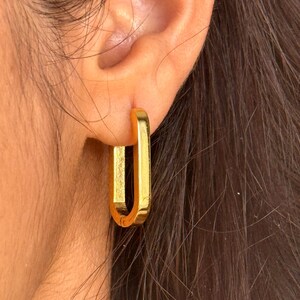 May include: A polished gold-tone hoop earring with a rectangular, elongated shape. The earring features a smooth, reflective metallic surface and a hinged closure mechanism, securely fastened through the earlobe with a clean, minimalist design.