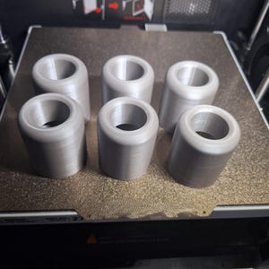 Peut inclure: Six objets cylindriques gris avec une base évasée sont disposés sur une surface texturée. Les objets ont un centre creux et semblent être imprimés en 3D. Les articles sont espacés uniformément et semblent être des composants ou des pièces.