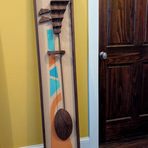 May include: Abstract wooden wall art in a rectangular frame. The artwork features a series of wooden shapes and a central, elongated oval. The background is a tan fabric with orange and teal geometric shapes. The frame is dark brown.
