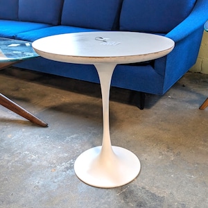 May include: White tulip table with a round top and a single pedestal base. The table is sitting on a concrete floor in front of a blue sofa.