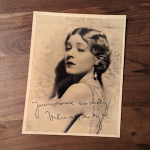 Puede incluir: Fotografía en blanco y negro de una mujer con el pelo corto y ondulado. Lleva un vestido de color claro y un collar de perlas. La mujer mira hacia un lado y sonríe ligeramente. La foto está firmada "Yours most sincerely, Vilma Banky".