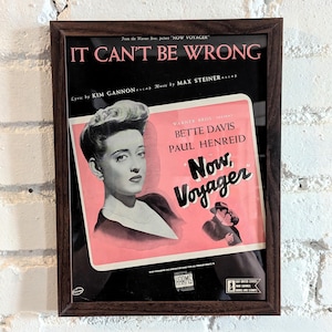 Op de afbeelding: Een ingelijste vintage filmposter voor de film "Now, Voyager" met Bette Davis en Paul Henreid. De poster heeft een roze achtergrond met de titel in grote witte letters en een zwart-witte afbeelding van Bette Davis. De tekst op de poster luidt "From the Warner Bros. picture "Now, Voyager" It Can't Be Wrong Lyric by Kim Gannon Music by Max Steiner."