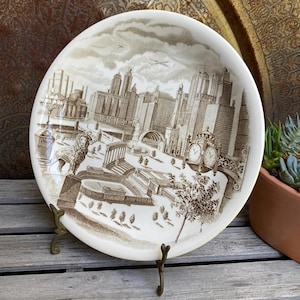 Puede incluir: Un plato decorativo con una ilustración en tonos sepia de un horizonte urbano, con edificios, una torre del reloj y un parque. El plato se exhibe sobre un soporte de latón, mostrando su diseño.