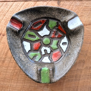 May include: Triangular ashtray with a textured, speckled gray exterior. The center features a stained-glass-like design with red, green, white, and gray segments. The ashtray has three cigarette rests, with red, white, and green accents.
