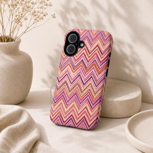 Op de afbeelding: Een smartphonehoesje met een kleurrijk zigzagpatroon in roze, paarse en oranje tinten. De hoes heeft een camera-uitsparing en zwarte knoppen aan de zijkant. Het ligt op een kleine, ronde, beige steen.