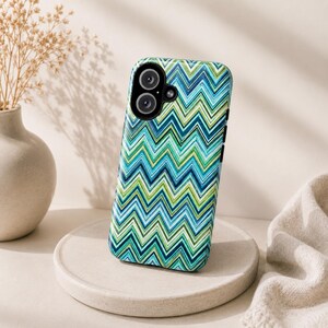 Op de afbeelding: Een smartphonehoesje met een levendig zigzagpatroon in blauw- en groentinten. De hoes heeft een glanzende afwerking en uitsparingen voor de camera en knoppen. De telefoonhoes wordt gepresenteerd op een ronde, neutraal gekleurde sokkel.