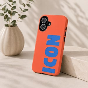 Peut inclure: Coque de téléphone orange avec le mot "ICON" en lettres bleues. L'étui présente un découpage pour un appareil photo à double objectif et des boutons noirs sur le côté. Un vase avec une plante est en arrière-plan.