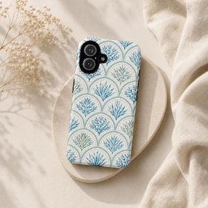 Op de afbeelding: Een smartphonehoesje met een herhalend patroon van gestileerde blauwe en groene plantmotieven. De hoes heeft een witte achtergrond, een cirkelvormige camera-uitsparing en een zwarte verticale streep. Het ligt op een beige, cirkelvormig oppervlak.