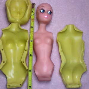 May include: A selection of doll-making components: a pink doll torso, a yellow head mould, and two yellow torso moulds. The doll torso is approximately 13 cm tall, as indicated by a measuring tape.