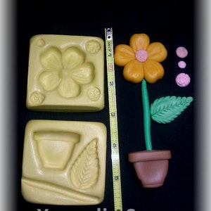 Puede incluir: Dos moldes de silicona para hacer decoraciones con forma de flor. Un molde tiene una sola flor con cinco pétalos, el otro tiene una maceta, una hoja y una flor pequeña. Los moldes son amarillos y las flores son naranja, rosa y verde.