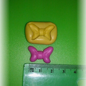 May include: Two bow-shaped ornaments. One is a light yellow color, rectangular, and has a bow design. The other is a small, vibrant pink bow. A green ruler with centimeter markings is in the foreground, showing the size of the bows.