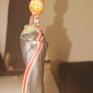 Könnte beinhalten: Geschnitzte Holzfigur einer Person, die ein gestreiftes orange-weißes Gefäß auf dem Kopf trägt. Die Figur ist in ein schwarzes Gewand mit roten und weißen Akzenten gehüllt. Die Figur steht auf einem hellbraunen Sockel.