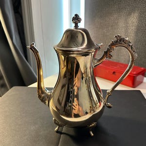 May include: A silver-colored teapot with a decorative handle and spout. The teapot has a lid with a small, ornate finial. The pot sits on three small feet. The polished surface reflects the surrounding environment.