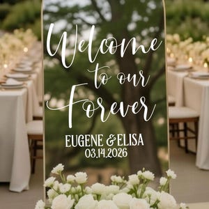 May include: An arched mirror wedding welcome sign with a gold frame, featuring white script text that reads Welcome to our Forever, followed by EUGENE & ELISA and the date 03.14.2026, decorated with a base arrangement of white roses and hydrangeas.
