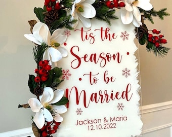 May include: White acrylic sign with red lettering that reads "'Tis the Season to be Married" with the names Jackson & Maria and the date 12.10.2022. The sign is decorated with a white magnolia flower garland with red berries and pine cones.