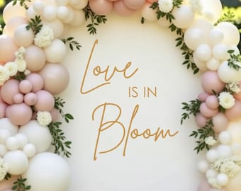 May include: An arched white backdrop featuring the gold text Love IS IN Bloom, decorated with a dense arrangement of white and soft pink balloons of varying sizes, interspersed with small white floral clusters and green leafy sprigs.