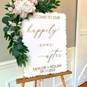 Peut inclure: Une pancarte en acrylique blanche avec des lettres dor&eacute;es qui disent "Bienvenue dans notre bonheur pour toujours Taylor + Nolan 09.17.2022". La pancarte est d&eacute;cor&eacute;e d'une guirlande de fleurs blanches et de feuilles vertes.