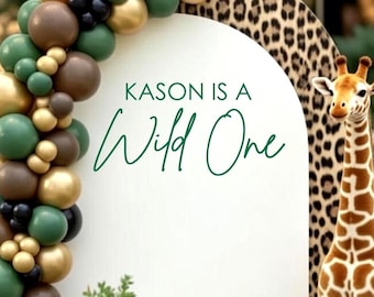 May include: An arched backdrop with a leopard print border and a white center featuring the text KASON IS A Wild One. A balloon garland in shades of green, gold, and brown curves along the left side, accompanied by a plush giraffe figure.