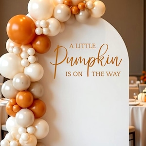 May include: A white arched backdrop featuring the text A LITTLE PUMPKIN IS ON THE WAY in gold lettering. A balloon garland in shades of orange, cream, and white is attached to the left side of the arch, cascading from the top to the floor.