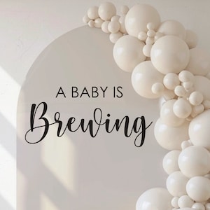 May include: A cream-colored arch with the text "A BABY IS Brewing" in black script. A cascade of white balloons adorns the right side. The image is a decal, not a backdrop, and will not stick to fabrics or spandex arch covers.
