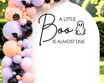 May include: A white arched backdrop featuring the black text A LITTLE Boo IS ALMOST DUE with a ghost icon. A balloon garland in shades of lavender, peach, and black is attached to the left side, accented with black paper bat and pumpkin cutouts.