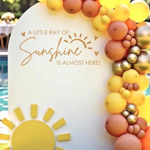 May include: A white, arched sign with a yellow sun design and the text "A LITTLE RAY OF SUNSHINE IS ALMOST HERE!" is decorated with a balloon arch in shades of yellow, orange, and gold. The sign is set up outdoors near a pool.