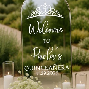 May include: A clear, arched mirror with the words "Welcome to Paola's Quinceañera 11.29.2025" in white script. The mirror is decorated with a crown design. White candles in glass cylinders and white flowers are arranged in front of the mirror.