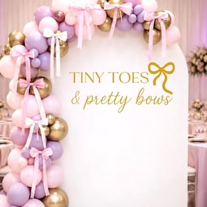 May include: A white arched backdrop featuring the gold text Tiny Toes & pretty bows with a gold bow icon. An asymmetrical balloon garland in shades of pink, lavender, and metallic gold accents the arch, decorated with several light pink ribbons.