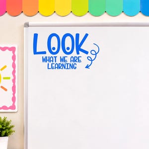 May include: A white dry-erase whiteboard featuring blue vinyl lettering that reads LOOK WHAT WE ARE LEARNING with a curly arrow graphic. The board has a silver metal frame and a tray at the bottom holding a row of colorful dry-erase markers.
