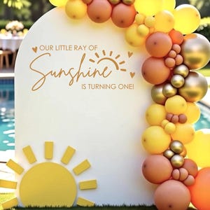 May include: A white, arched sign with the text "OUR LITTLE RAY OF SUNSHINE IS TURNING ONE!" is decorated with a yellow sun and a balloon arch in shades of yellow, orange, and gold. The sign is set up outdoors near a pool.