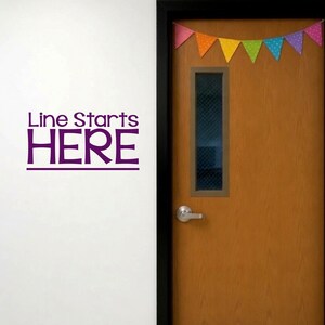May include: A white wall featuring a purple vinyl decal that reads Line Starts HERE with a horizontal line underneath. To the right is a wooden door with a rectangular window, a silver handle, and a string of colorful polka dot bunting flags.