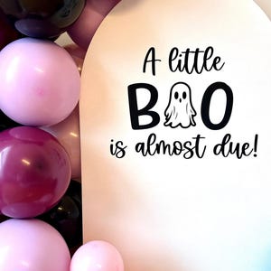 May include: A festive balloon arch in shades of pink, burgundy, and black surrounds a white sign. The sign reads "A little BOO is almost due!" in black script, with a ghost graphic.