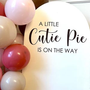 May include: A white arched sign with black text that reads A LITTLE Cutie Pie IS ON THE WAY, accompanied by a cluster of balloons in shades of pink, dark brown, and cream. The sign features a smooth surface and a rounded top edge.