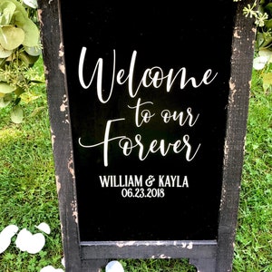 May include: A black chalkboard sign with a distressed wooden frame, decorated with green eucalyptus garlands. The sign features white text that reads Welcome to our Forever, WILLIAM & KAYLA, and the date 06.23.2018 in a mix of script and sans-serif fonts.