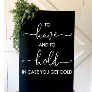 May include: A rectangular black sign featuring white text that reads TO have AND TO hold IN CASE YOU GET COLD in a mix of script and sans-serif fonts. The sign is decorated with a sprig of green eucalyptus leaves attached to the top left corner.
