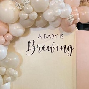 May include: A baby shower backdrop featuring a balloon arch in shades of white and blush pink. The arch frames a wooden panel with the phrase "A Baby Is Brewing" in elegant script. The backdrop is set against a backdrop of wooden barrels.