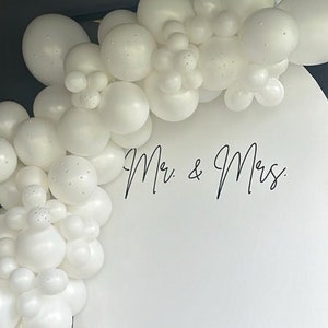 May include: White balloon arch with the words "Mr. & Mrs." on a white backdrop. The balloons are arranged in a cascading pattern, creating a festive and romantic atmosphere.