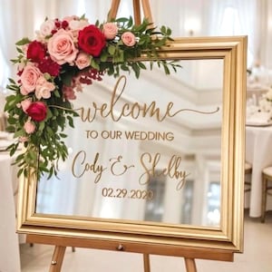 Może przedstawiać: Prostokątne lustro w złotej ramie umieszczone na drewnianej sztaludze. Lustro posiada złoty napis "welcome TO OUR WEDDING Cody & Shelby 02.29.2020" i jest ozdobione bukietem czerwonych oraz r&oacute;żowych r&oacute;ż wraz z zielonymi liśćmi.