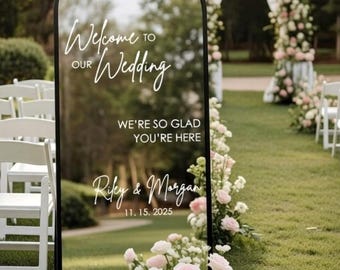 May include: An arched mirror wedding sign with a black frame featuring white text that reads Welcome to our Wedding, We're so glad you're here, Riley & Morgan, and 11. 15. 2025. The mirror reflects the surrounding outdoor garden setting.