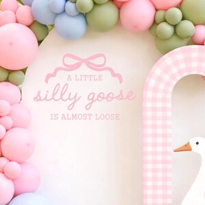 May include: A whimsical party decoration featuring a white arch with a pink and white gingham archway and a large white goose cutout. The arch is adorned with a balloon garland in pastel pink, blue, and green. Text reads "A little silly goose is almost loose."