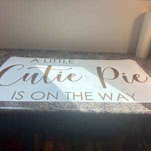 May include: A rectangular white vinyl decal featuring the brown text A LITTLE Cutie Pie IS ON THE WAY. The words A LITTLE and IS ON THE WAY are in a simple sans-serif font, while the words Cutie Pie are in a larger, elegant script font.