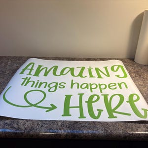 Puede incluir: Una hoja de vinilo adhesivo blanca con el texto verde "Amazing things happen HERE" en una mezcla de fuentes script y sans-serif. El dise&ntilde;o incluye una flecha decorativa que forma un bucle bajo la palabra "Amazing" y apunta hacia "HERE".