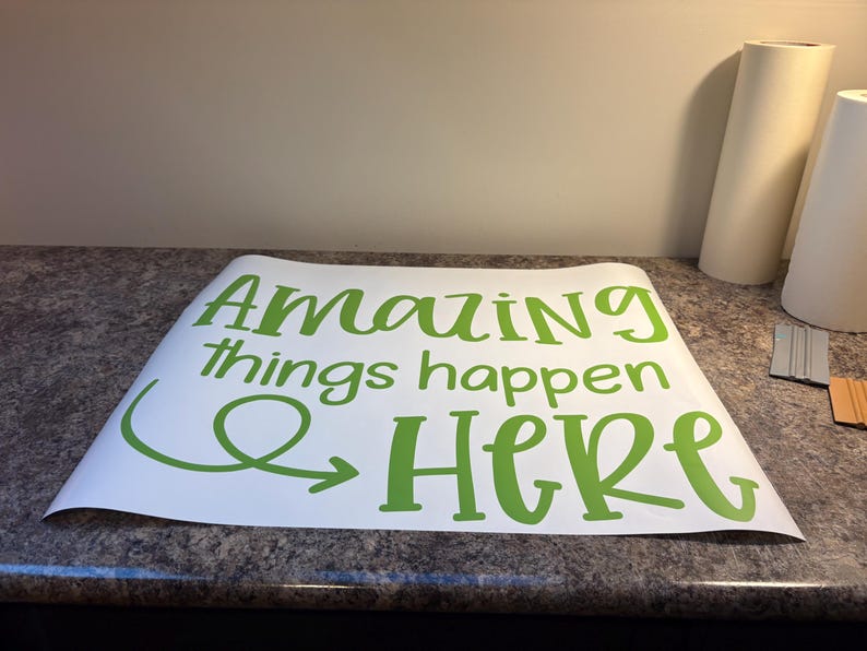 Puede incluir: Una hoja de vinilo adhesivo blanca con el texto verde "Amazing things happen HERE" en una mezcla de fuentes script y sans-serif. El dise&ntilde;o incluye una flecha decorativa que forma un bucle bajo la palabra "Amazing" y apunta hacia "HERE".