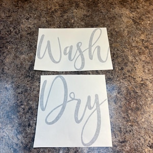 Puede incluir: Dos calcoman&iacute;as rectangulares blancas con texto gris en cursiva. La calcoman&iacute;a superior dice "Wash", y la calcoman&iacute;a inferior dice "Dry". Las calcoman&iacute;as se muestran sobre una superficie marr&oacute;n moteada.